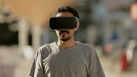 Junger-Arabischer-Mann-Mit-Dunklem-Lockigem-Haar-Und-Bart-Im-Grauen-T-Shirt,-Der-Mit-Einer-Virtual-Reality-Brille-Im-Park-Ist